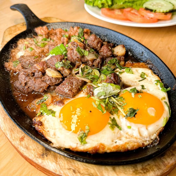 Vietnamese sizzling steak and eggs (Bò Né) in a cast iron skillet, garnished with herbs.