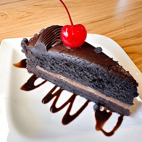 Slice of rich chocolate cake with chocolate frosting, a red cherry, and chocolate drizzle on a white plate.