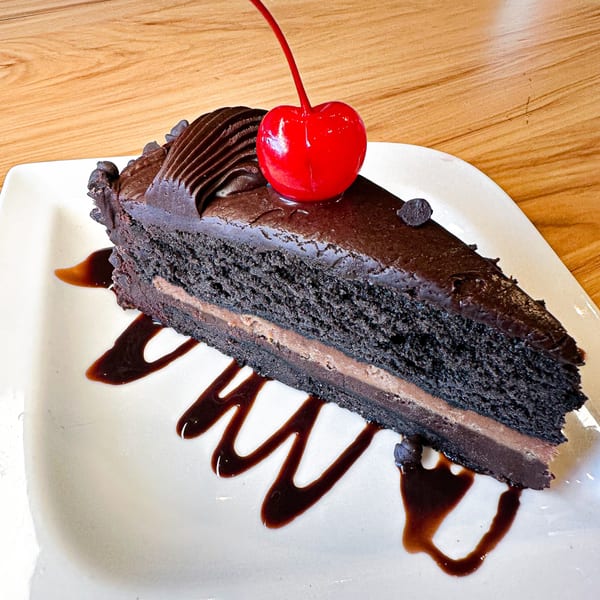 A slice of rich chocolate cake with chocolate frosting, a red cherry, and chocolate drizzle.